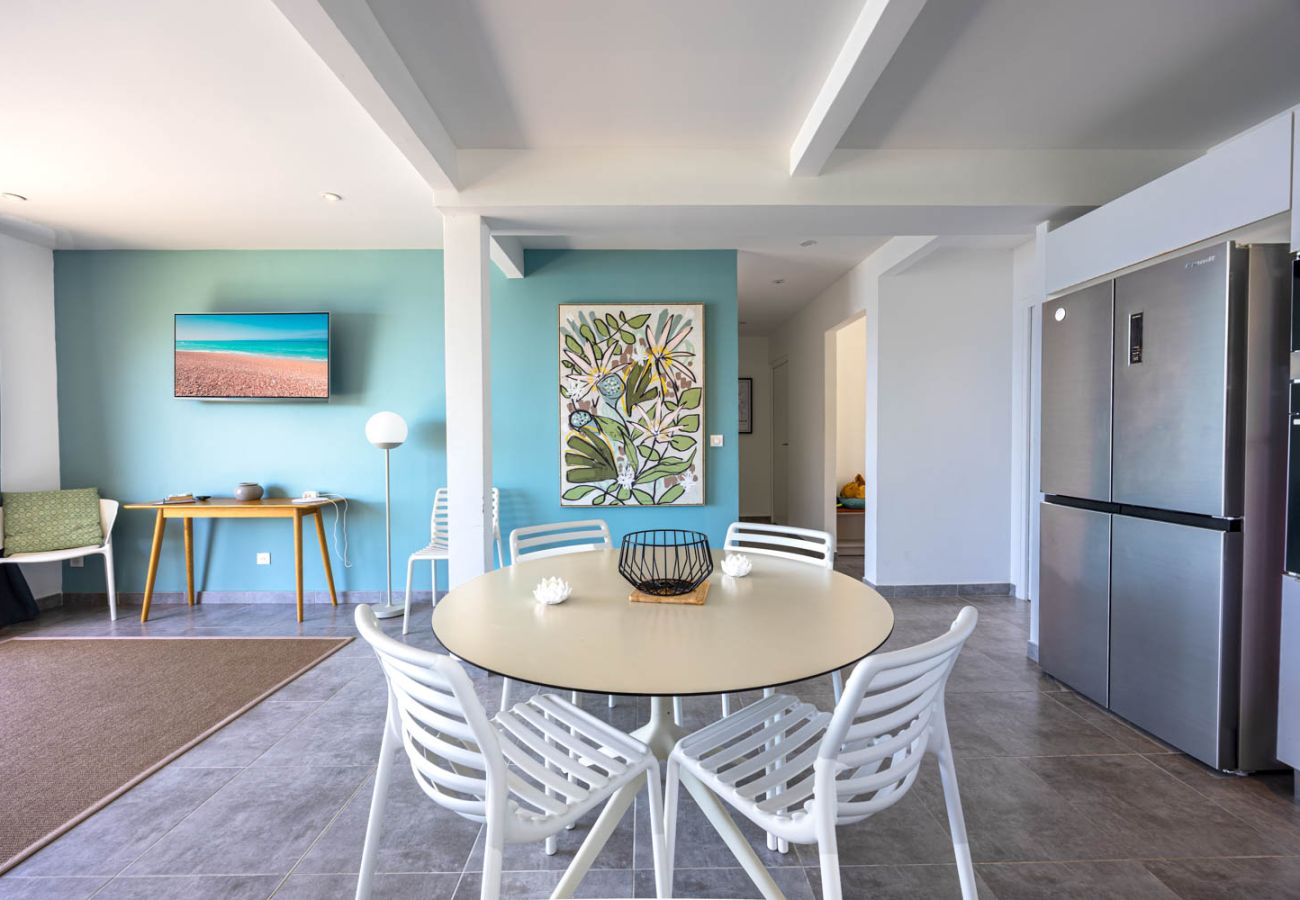 Air-conditioned living room with blue wall, white round table and modern fitted kitchen at Villa Ti Soleil in Anses-d'Arlet.