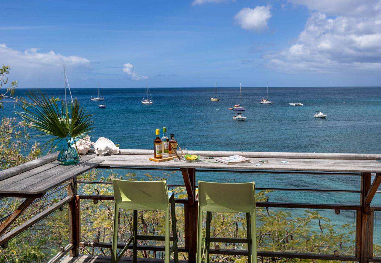 Comptoir de bar en bois sur une terrasse, deux tabourets verts et vue panoramique sur les voiliers aux Anses-d'Arlet.